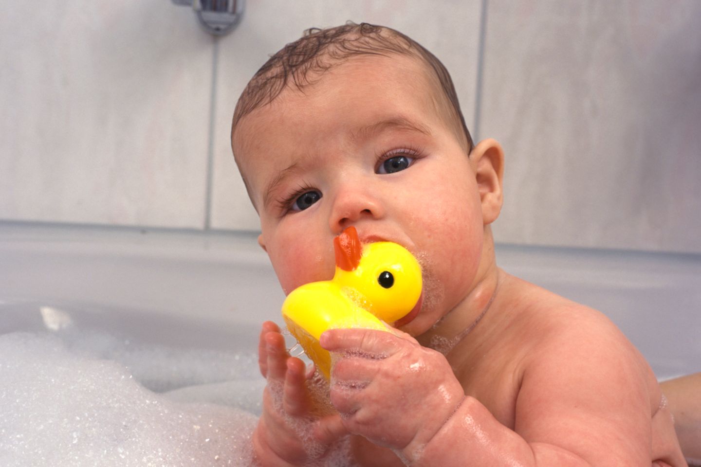 Baby sitzt in Badewanne und lutscht an Babdeente