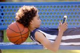 Junge liegt auf einer Bank,mit dem Kopf auf einem Basketball, hat ein Handy in der Hand