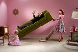 Fastenzeit: Frau hebt ein Sofa hoch auf dem ein Mann liegt und saugt dabei Staub