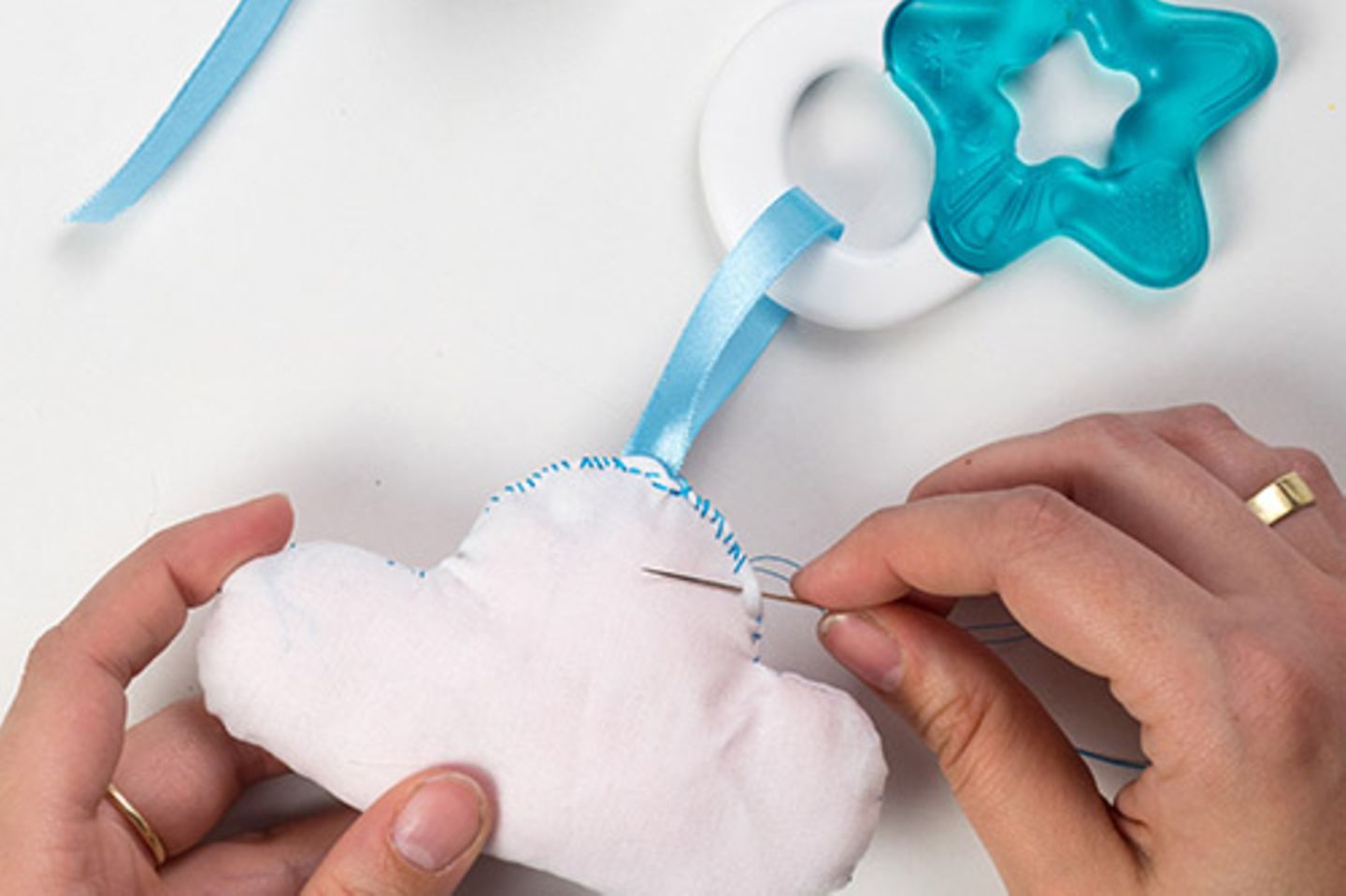 Ein Stück Satinband (10 bis 12 cm) abschneiden, durch den Beißring ziehen und die Enden zusammen in die Öffnung der Stoff gur stecken. Die offenen Kanten nach innen schlagen und alles mit einer Nadel sichern. Nun die offene Kante zunähen, dabei das Satinband mit einnähen.    