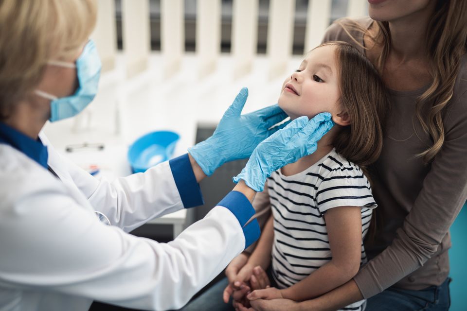 Ein Mädchen sitzt auf dem Schoß der Mutter und wird von einer Kinderärztin mit Schutzkleidung untersucht