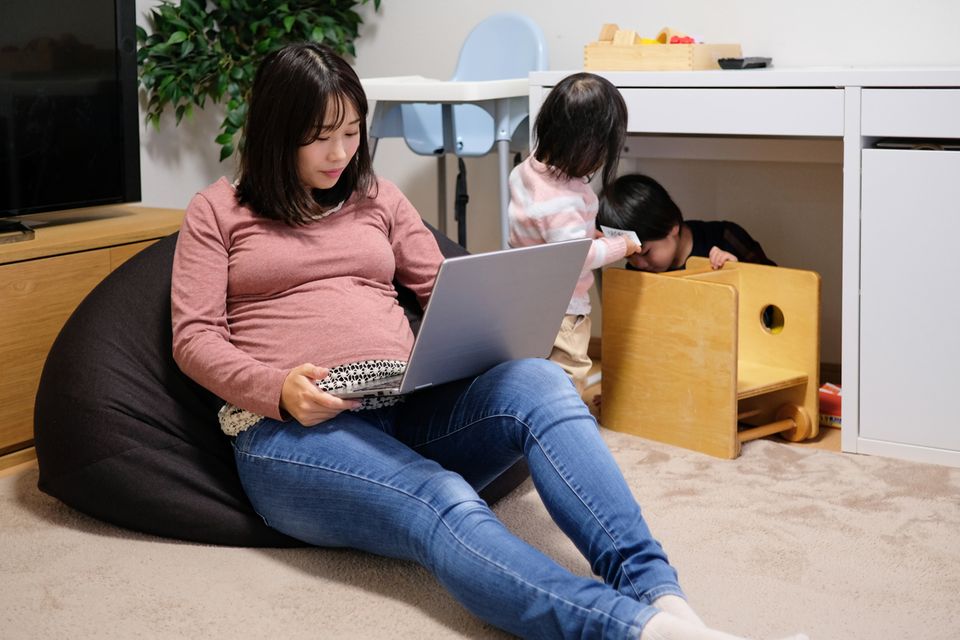Schwangere Mama am Laptop mit zwei Kindern im Hintergrund