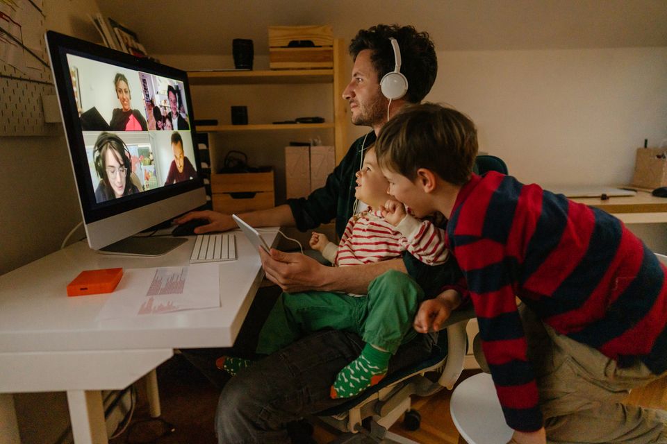 Ein Vater befindet sich in einer Videokonferenz, seine beiden Kinder schauen zu