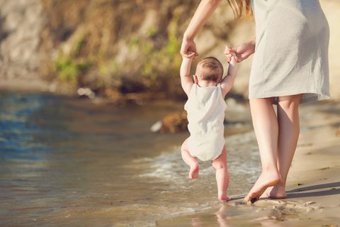 Mutter geht mit Baby an der Hand am Meer spazieren