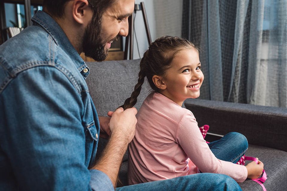 Vater flicht Tochter einen Zopf auf dem Sofa.