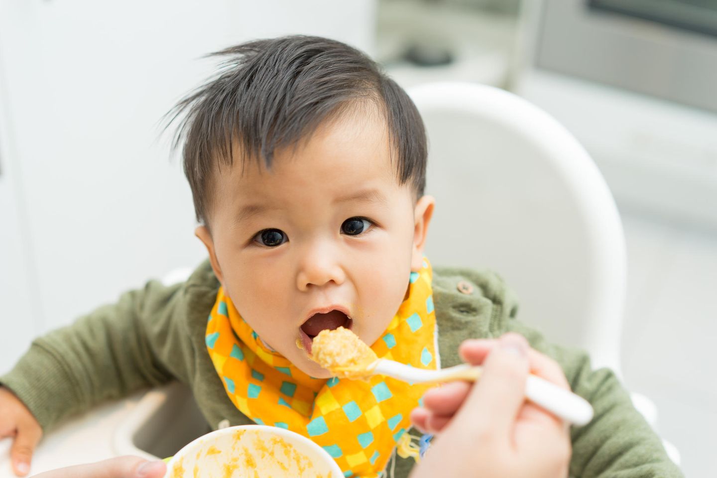 Beikost einführen: Asiatisches Baby sitzt im Hochstuhl und wird mit einem Löffel Beikost gefüttert
