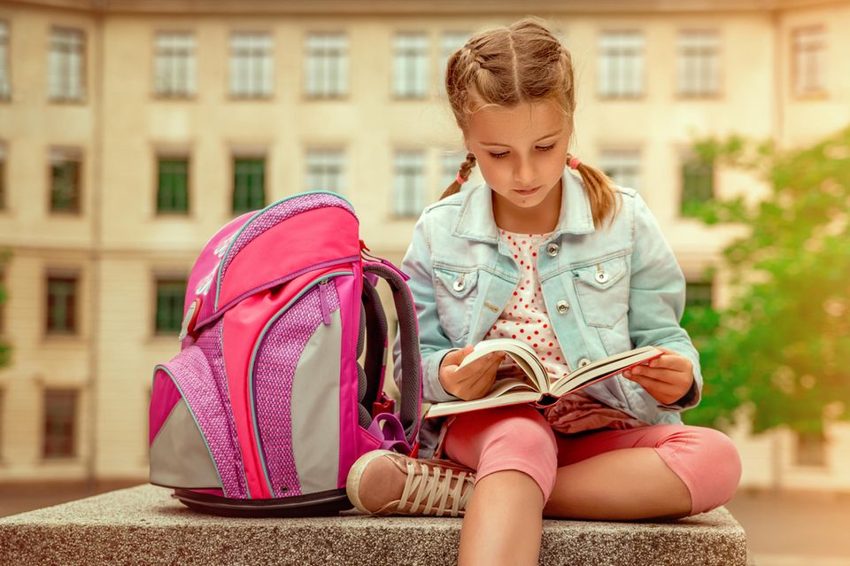 Schulranzen-Test: Mädchen mit rosa Schulranzen neben sich blättert im Buch vor der Schule.