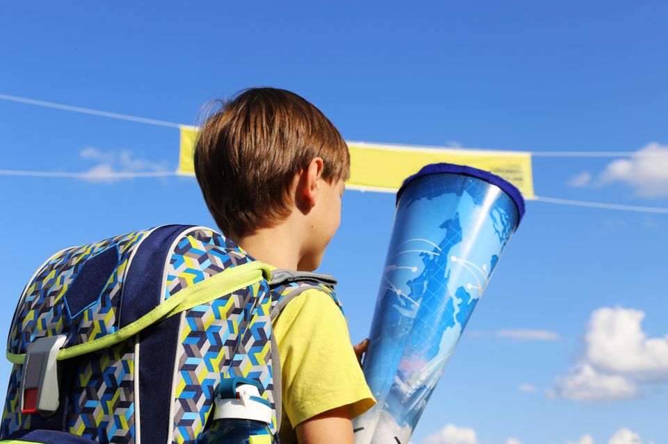 Einschulung: Junge mit Brille sitzt mit Schultüte und Schulranzen auf dem Schulhof.