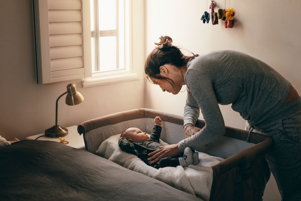 Junge Mutter beugt sich über Beistellbett um ihr Baby zu beruhigen.