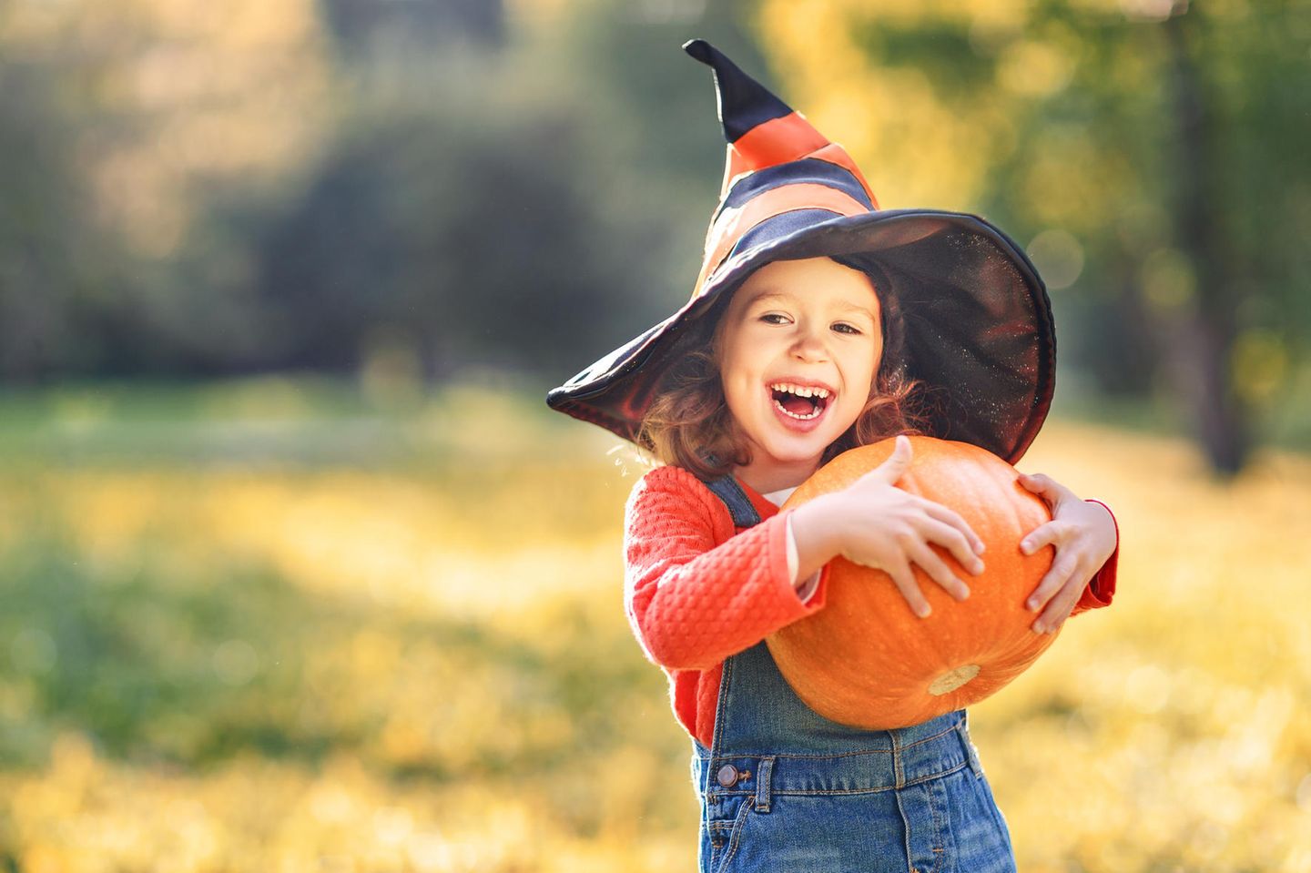 Mädchen mit Hexen-Hut hält einen Halloween Kürbis und lacht