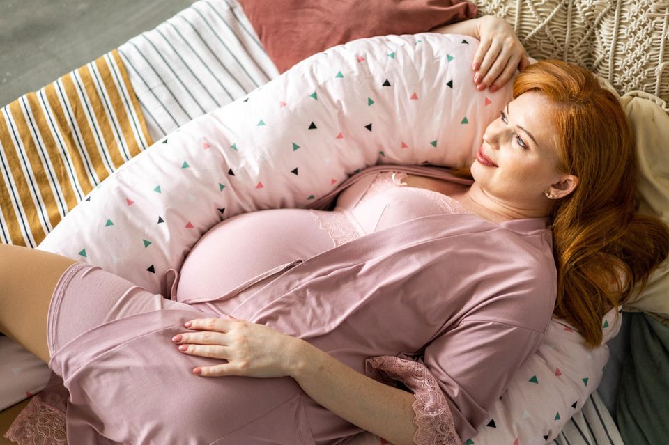 Wehen fördern: Hochschwangere Frau liegt verträumt mit einem Stillkissen im Bett