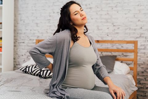 Eine schwangere Frau sitzt auf dem Bett und hält sich mit schmerzverzerrtem Gesicht den Rücken