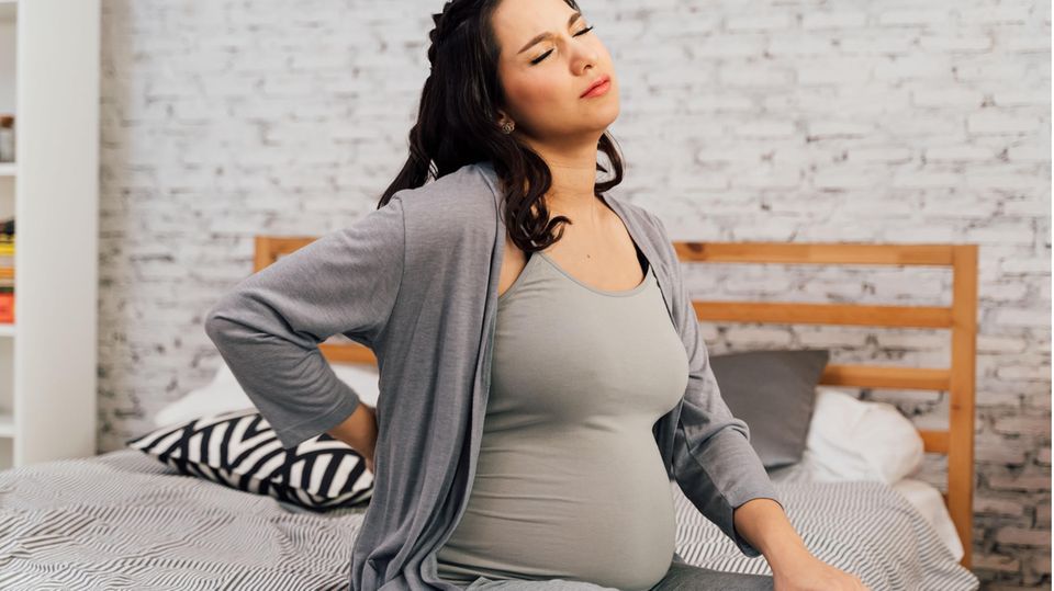 Eine schwangere Frau sitzt auf dem Bett und hält sich mit schmerzverzerrtem Gesicht den Rücken