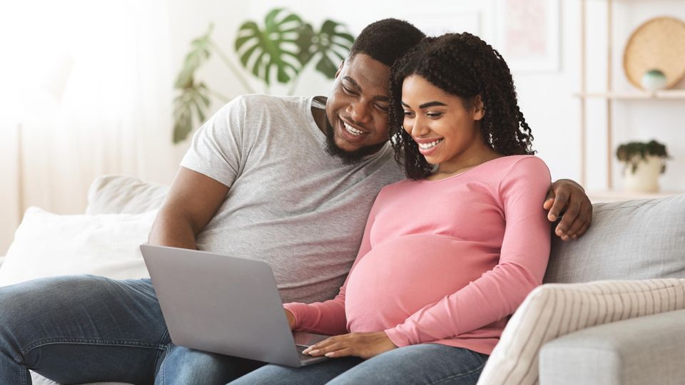 Ein Mann und eine schwangere Frau sitzen auf dem Sofa vor dem Laptop.