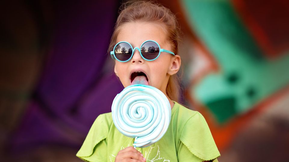 Ein Mädchen mit einer blauen Sonnenbrille auf der Nase lutscht an einem großen blauen Lolli.