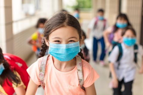 Ein Mädchen mit Mund-Nasen-Schutz läuft auf einem Schulgang mit ihren Mitschülern und lacht in die Kamera.