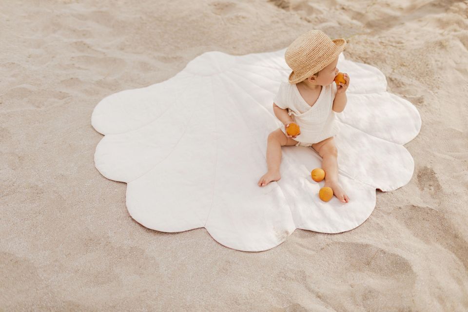 Umweltfreundlich: Baby sitzt auf einer Decke im Sand und hat Orangen in der Hand