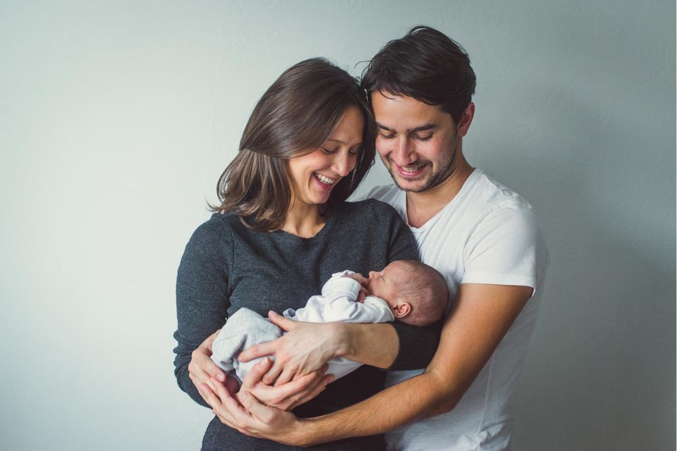 ein junges Elternpaar hält ein Neugeborenes in den Armen und lächelt es an.