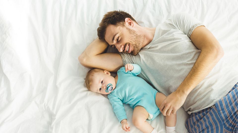 Ein Mann und ein Baby mit Schnuller liegen im Bett.