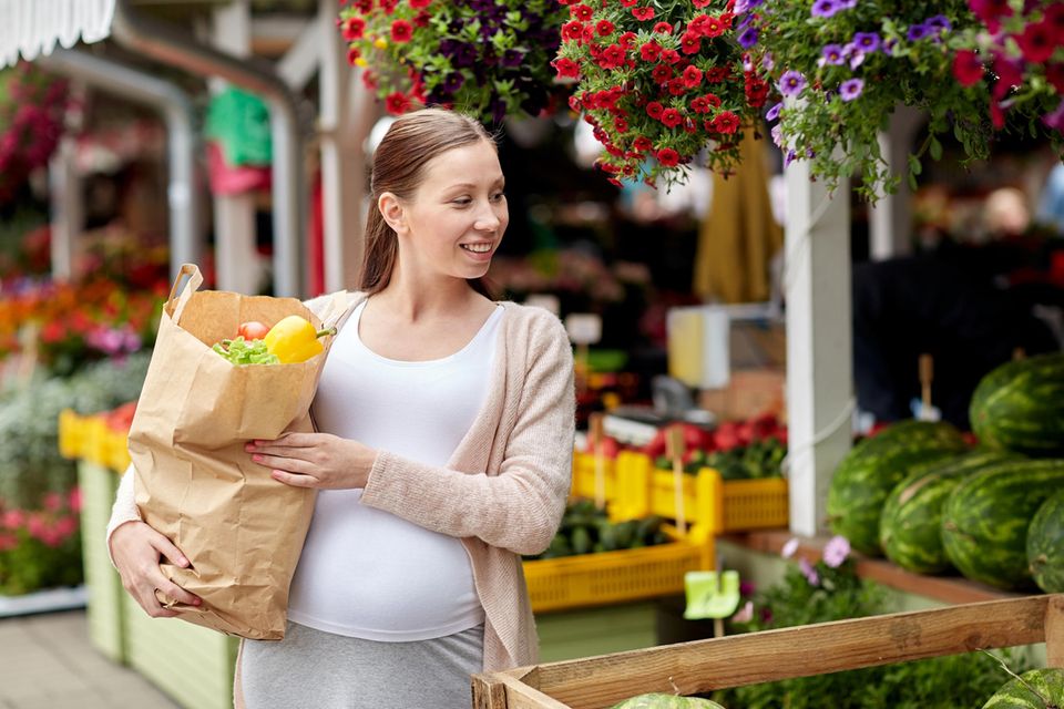 Eine schwangere Frau ist auf einem Wochenmarkt einkaufen und trägt eine Tüte mit Gemüse auf dem Arm
