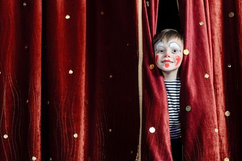 Clown schminken: Ein als Clown geschminkter Junge steht in schwarz-weißem T-Shirt hinter einem roten Vorhang und grinst