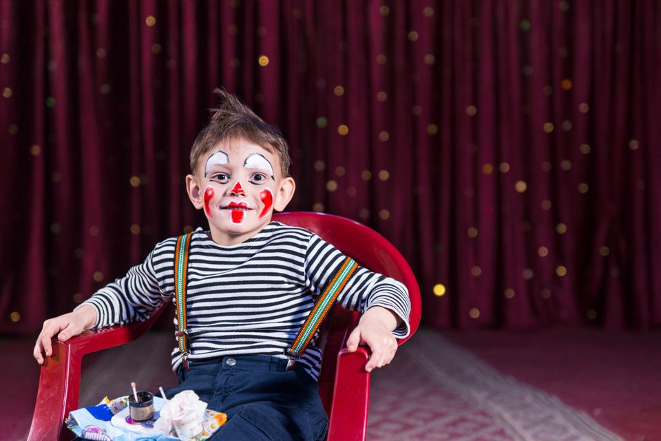 Clown schminken: Ein Junge sitzt auf einem roten Stuhl und hat ein geschminktes Clownsgesicht