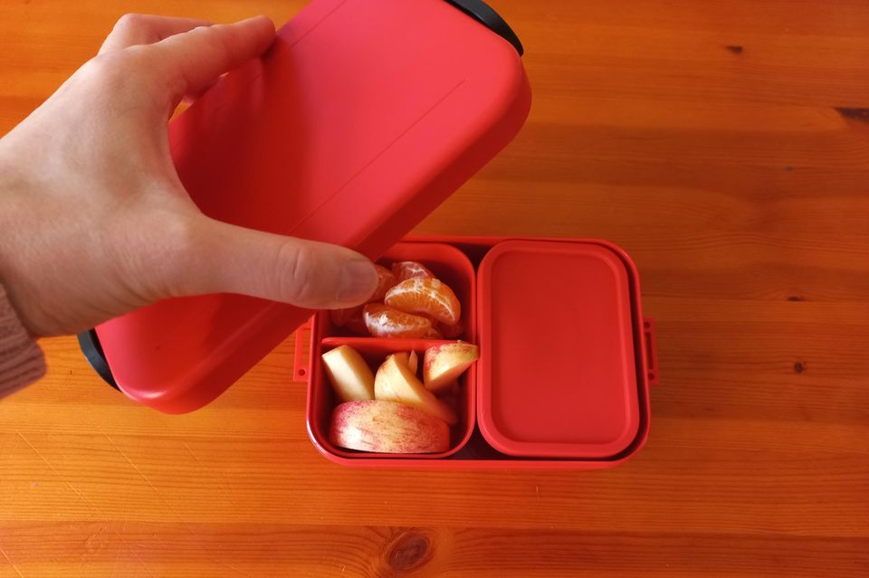 Hand hält den Deckel über einer roten Bento-Box auf dem Küchentisch.