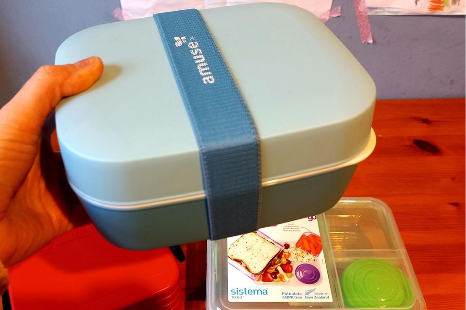 Hand hält eine hellblaue Bento-Box vor einem Küchentisch mit anderen Bento-Boxen.