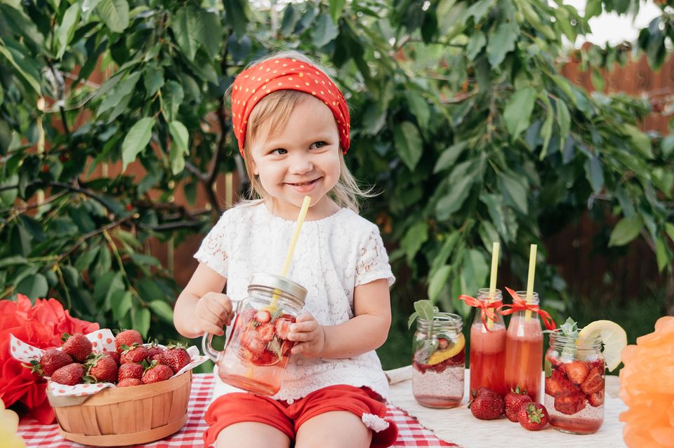 Kinderbowle selbst machen: Blondes Mädchen sitzt auf einem Tisch, in der Hand ein Glas mit Erdbeerbowle