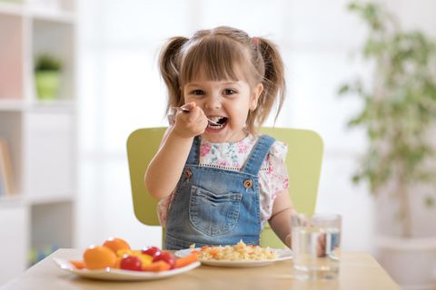 Ein kleines Mädchen sitzt auf einem grünen Stuhl am Tisch und isst lächelnd Reis