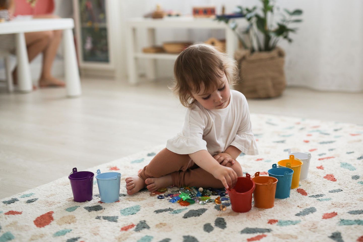 Montessori-Spielzeug: Mädchen auf dem Teppich sortiert Glassteinchen in bunte Becher.