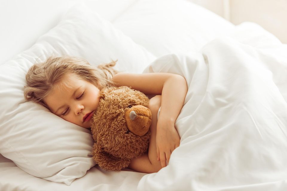 Bettdecke im Test: Blondes Mädchen mit Teddybär schläft im Bett unter weißer Bettwäsche.
