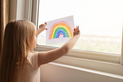 Ein Kind steht am Fenster und hält ein Bild mit einem Regenbogen hoch.