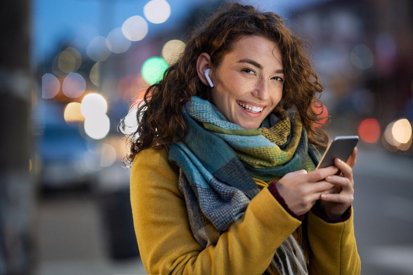 Lachende junge Frau hört abends auf der Straße einen Postcast über Kopfhörer