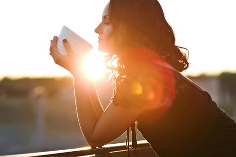Selbstfürsorge: ein Frau lehnt bei Sonnenaufgang gegen ein Geländer mit einem Becher in der Hand