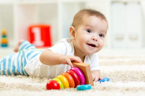 Ein Baby liegt auf dem Teppich und spielt mit buntem Spielzeug aus Holz