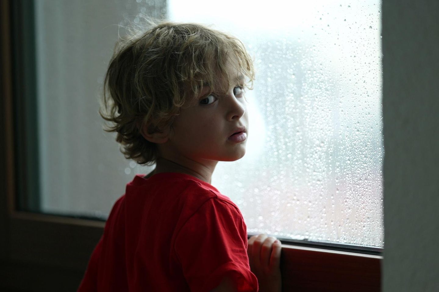 Psychische Beschwerden bei Kindern: ein kleinere blonder Junge steht am Fenster, es regnet