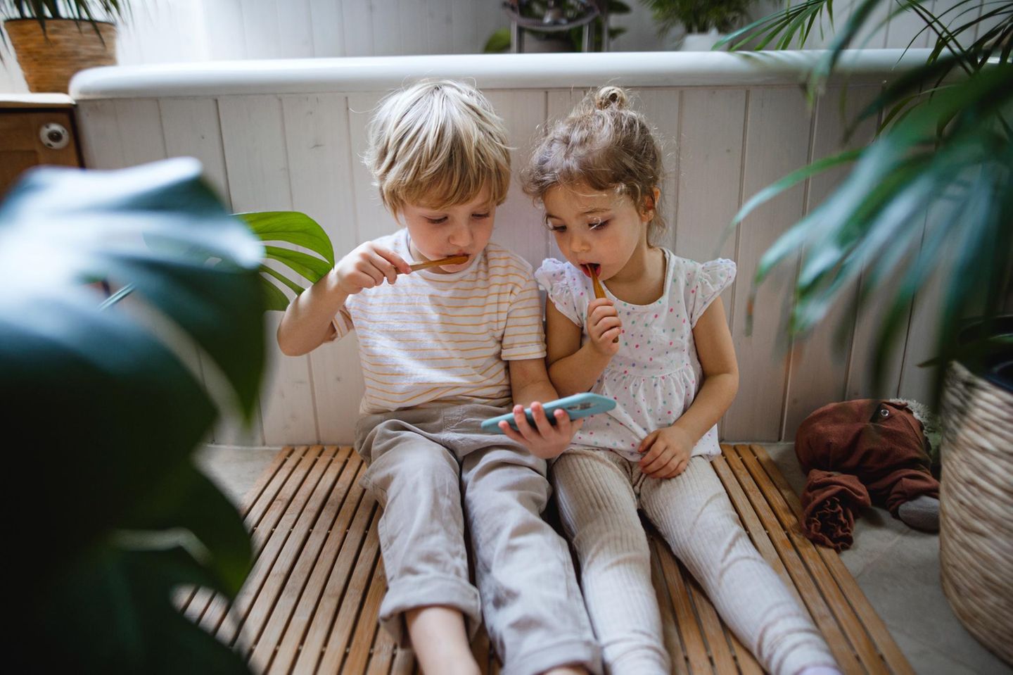 Smartphone fürs Kind: 2 kleine Kinder sitzen beim Zähneputzen mit einem Smartphone auf dem Boden