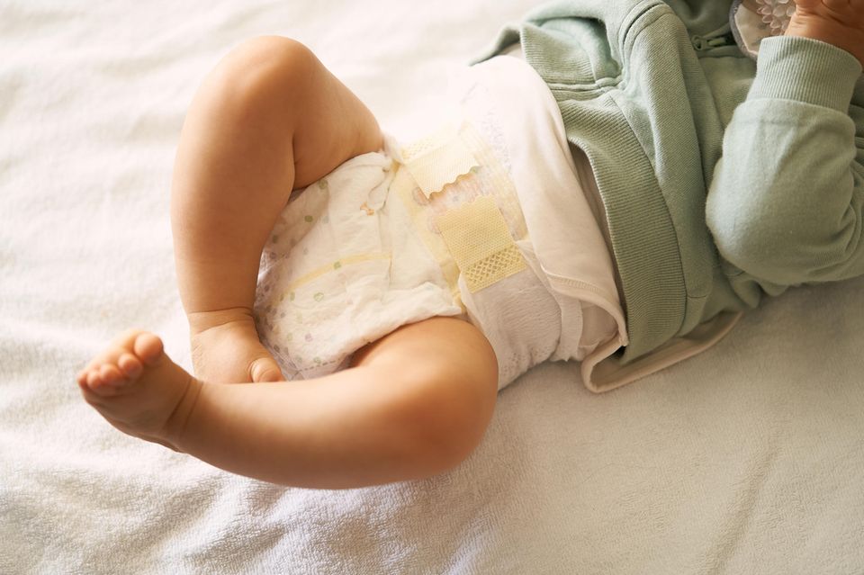 Windel-Test: Strampelndes Baby in Einwegwindel und Sweatshirt liegt auf dem Bett.