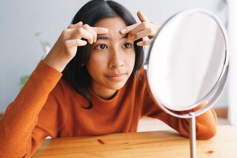 Pickel bei Kindern: ein asiatisches Mädchen betastet vor dem Spiegel ihr Gesicht