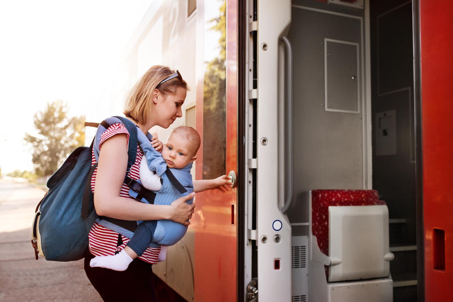 Reisen mit Baby: Junge Mutter mit Baby in der Trage und Rucksack steigt in einen Zug