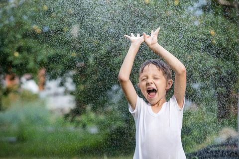 Wasserspielzeug: Junge im weißen T-Shirt erfrischt sich unter Wassersprinkler.