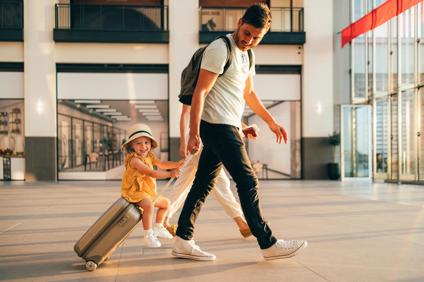 Flughafen-Chaos: Mädchen ist mit ihren Eltern am Flughafen. Sie sitzt auf dem Handgepäckkoffer und lacht.
