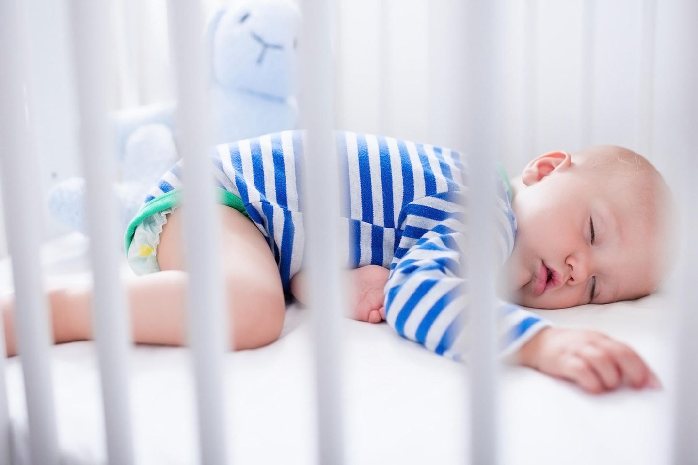 Baby im blau-weiß gestreiften Strampler schäft im Bett auf dem Bauch