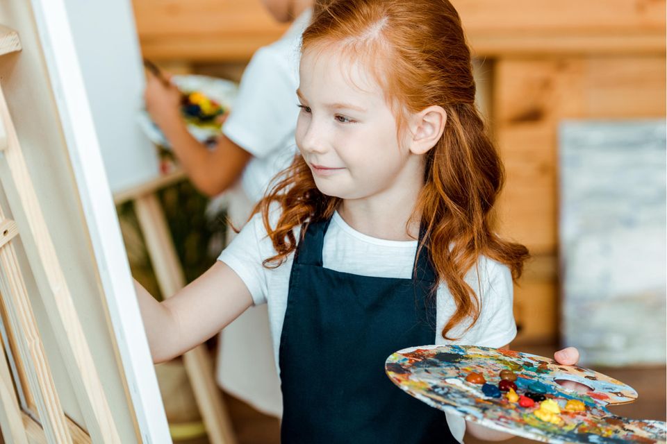 Malkittel für Kinder im Test: Mädchen mit Schürze und Farbpalette malt auf Leinwand.