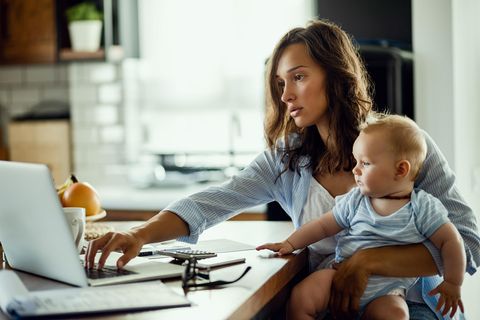Frau mit Säugling auf dem Arm sitzt vor dem Laptop