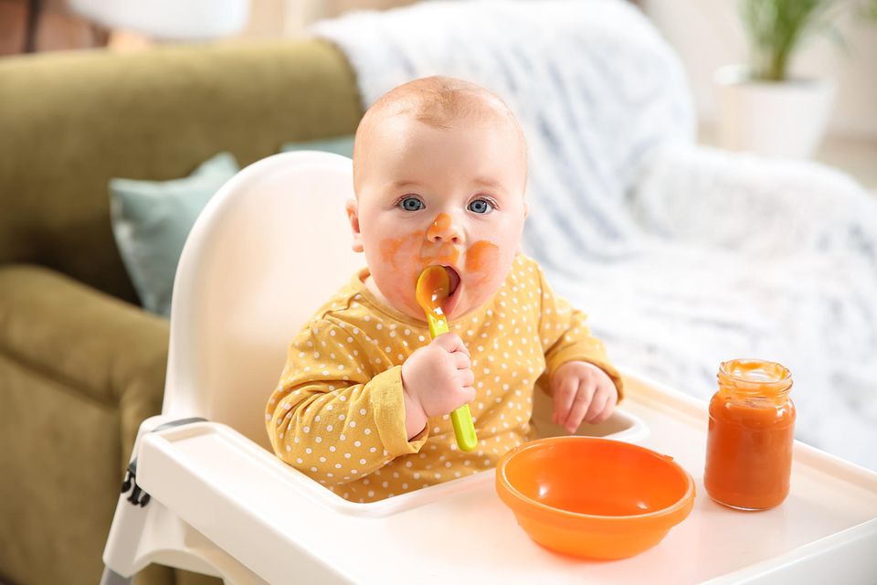 Experten empfehlen Essanfänger: ein Baby im gelben Body sitzt in einem Hochstuhl und isst orangenen Brei