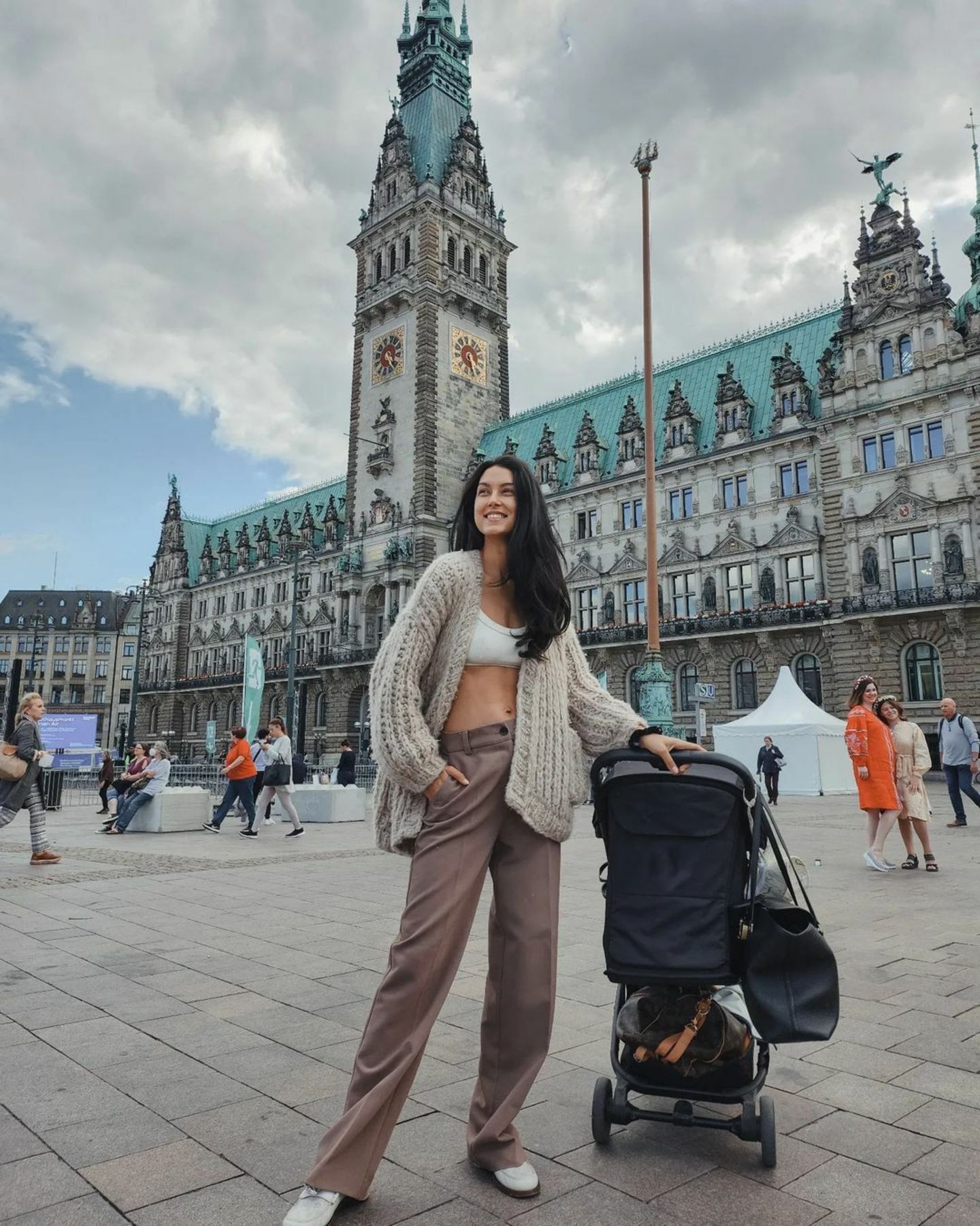 Mit Kinderwagen ist Rebecca Mir im September 2022 auf den Straßen Hamburgs unterwegs. Das deutsche Model trägt eine trendy Oversize-Strickjacke über einem weißen Brustiertop, dazu eine braune Stoffhose. 