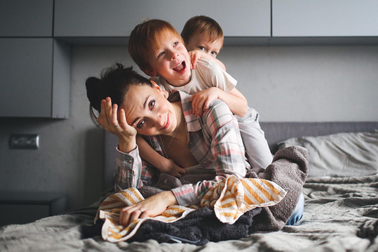 Erziehungsratgeber: Zweifelnde Mutter sitzt mit zwei tobenden Kindern auf dem Bett.