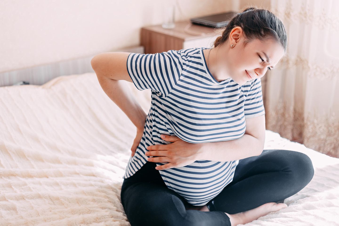 Schwangere sitzt mit schmerzverzerrtem Gesicht auf dem Bett und hält sich Rücken und Bauch